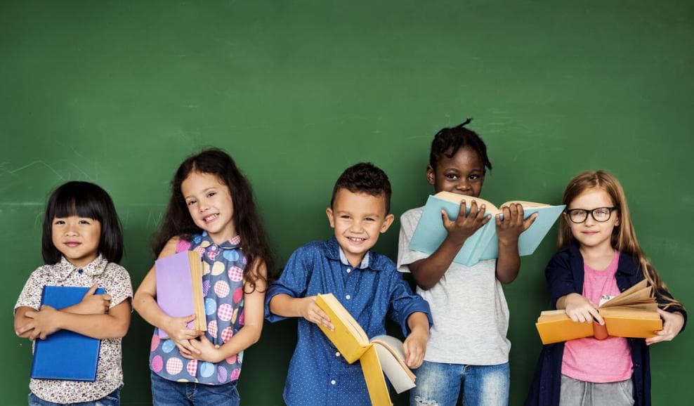 Five diverse elementary students holding books in front of a green chalkboard